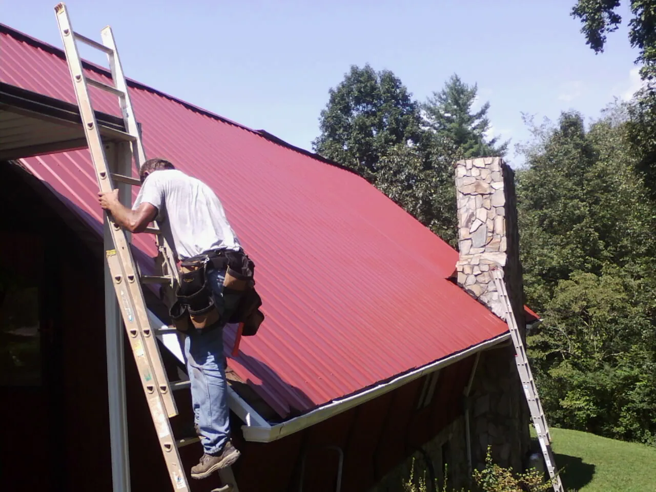 Roofer on a ladder beside a red metal roof and stone chimney — Soffit & Fascia Repair by Roofing Brothers in Kerby